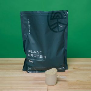a close-up of a dark green bag of Cymbiotika's Plant Protein supplement on a white oak surface against a bright green background, with a scoop of the powder sitting in front of it