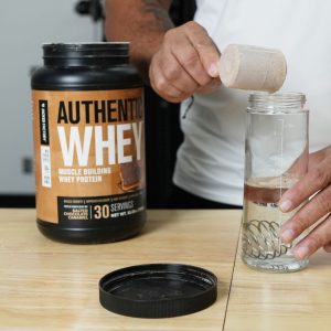 a person pouring a scoop of jacked factory authentic whey protein shake into a cup with water