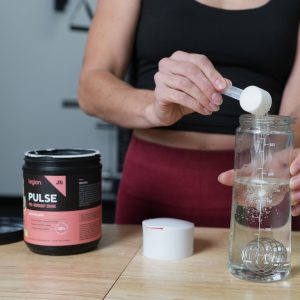 a young woman in a gym pouring a scoop of Legion Pulse pre-workout powder into a bottle of water