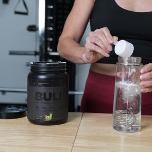 a young woman in a gym pouring a scoop of Transparent Labs Bulk Black preworkout powder into a bottle of water
