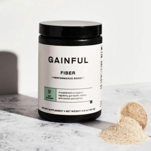 A container of Gainful Fiber Performance Boost supplement, with some powder spilled on the surface next to it. The label mentions gut support and performance boost.