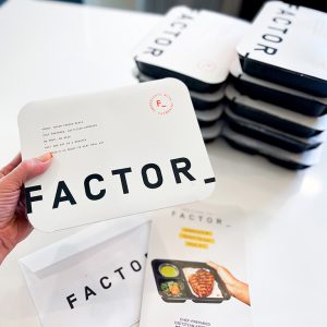 Ten prepared meals in their packaging from Factor on a white countertop, with one meal being held by the customer