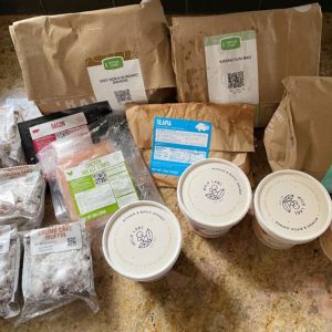 an assortment of delivered ingredients from Green Chef's meal kit, including tilapia, chicken breast strips, bacon, crumb cake muffins, and more on a countertop