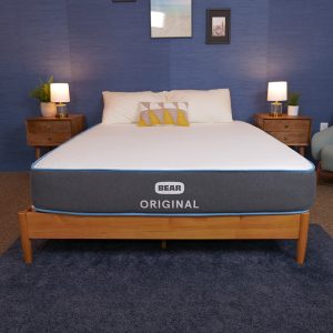 A bed with a gray mattress featuring a light blue stripe and a white top surface, set in a bedroom with a navy blue wall and two wooden nightstands with lamps. The bed is neatly made with pillows.