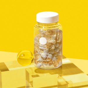 Clear bottle with a white cap labeled "Ritual Essential for Men 50+" containing clear capsules with small white beads inside, set against a yellow background.