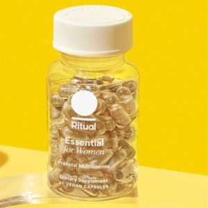 Bottle of Ritual Essential Prenatal Multivitamin with a white cap and transparent label, showing clear capsules, placed against a yellow background.