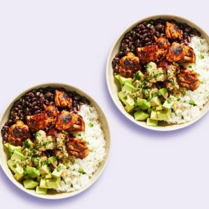 Two bowls from Dinnerly meal services, containing rice, black beans, avocado, and seasoned grilled chicken, garnished with herbs.