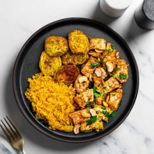 A black plate with roasted potatoes, yellow rice, and chicken pieces garnished with cashews and herbs.