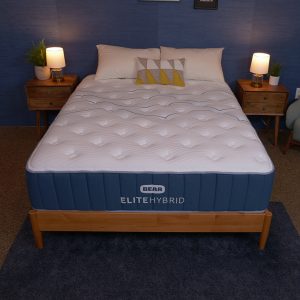 A bed with a dark blue mattress and a white quilted top surface, set in a bedroom with a navy blue wall and two wooden nightstands with lamps. The bed is neatly made with pillows.
